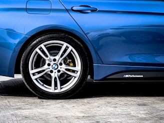 A close-up of a car's rear side, featuring a shiny, blue paint finish with a visible tire and sleek alloy wheel. The image captures the detailed design of the wheel and the M Performance badge on the side of the vehicle.