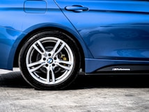 A close-up of a car's rear side, featuring a shiny, blue paint finish with a visible tire and sleek alloy wheel. The image captures the detailed design of the wheel and the M Performance badge on the side of the vehicle.
