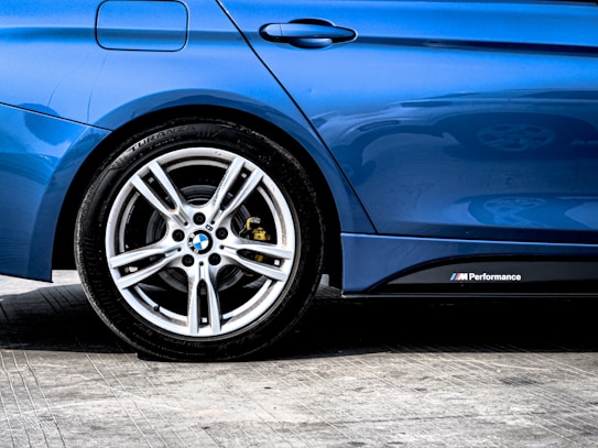 A close-up of a car's rear side, featuring a shiny, blue paint finish with a visible tire and sleek alloy wheel. The image captures the detailed design of the wheel and the M Performance badge on the side of the vehicle.