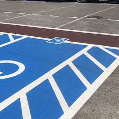 A parking area is painted with blue and white lines, featuring a sign indicating a reserved space. Cars are parked in adjacent spaces marked by yellow and white lines.