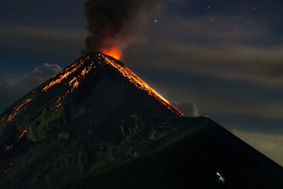 A dark, towering volcano erupts with red-hot lava flows streaming down its slopes, against a backdrop of a night sky with faint stars. Thick smoke and ash plume dramatically into the air from the fiery crater.