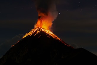 A volcanic eruption with bright orange lava spewing from the top, set against a dark night sky. Thick clouds of ash and smoke billow upwards, adding to the dramatic scene.