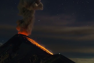 A volcano actively erupting at night with glowing lava streaming down its slopes and a thick plume of smoke rising into the starry sky.