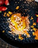 A vibrant culinary creation is arranged on a textured black plate. A yellow dessert, possibly a gourmet ice cream bar, is coated with a glossy layer and sprinkled with pine nuts. Surrounding the dessert are crumbles and scattered edible yellow flowers. Fresh strawberries are positioned artistically nearby.