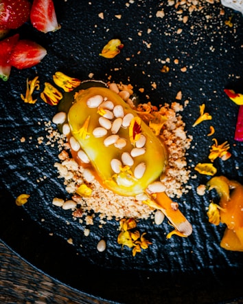 A vibrant culinary creation is arranged on a textured black plate. A yellow dessert, possibly a gourmet ice cream bar, is coated with a glossy layer and sprinkled with pine nuts. Surrounding the dessert are crumbles and scattered edible yellow flowers. Fresh strawberries are positioned artistically nearby.