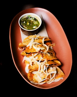 A plate with a series of folded, crispy tacos garnished with sliced onions, accompanied by a small bowl of green salsa. The colors are vibrant and the food arrangement is appetizing, presented on a warm, oval-shaped ceramic dish against a dark background.