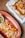 Crispy, golden-brown tacos with shredded onions spread across a red serving dish, accompanied by a small bowl of green salsa. A larger basket covered with a cloth contains more tacos, creating an inviting and rustic dining setting.