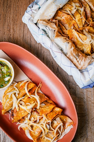 Crispy, golden-brown tacos with shredded onions spread across a red serving dish, accompanied by a small bowl of green salsa. A larger basket covered with a cloth contains more tacos, creating an inviting and rustic dining setting.