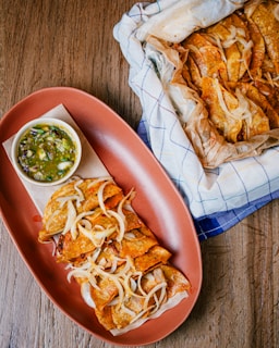 A wooden table features a platter of golden-brown fried tacos topped with grilled onions, accompanied by a small bowl of green salsa. Next to the platter is a parchment-lined basket filled with more tacos.