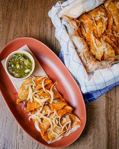 A wooden table features a platter of golden-brown fried tacos topped with grilled onions, accompanied by a small bowl of green salsa. Next to the platter is a parchment-lined basket filled with more tacos.