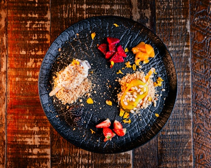 An artful arrangement of assorted desserts on a textured black plate, featuring a variety of colors and textures. There are two small dessert items, one covered in crumbs with white icing and another topped with a yellow glaze and white seeds. Additional elements include pieces of vibrant red fruit, possibly strawberries, and slices of another red and yellow fruit.