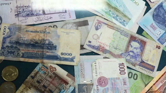 A collection of various international currencies, including notes and coins arranged on a surface. The banknotes feature intricate designs, different scripts, and diverse colors, reflecting the unique characteristics of each country's currency.