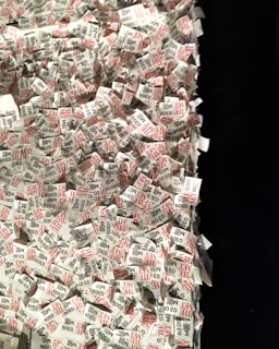 A large pile of printed paper tags, predominantly featuring printed text in black and red. Each tag includes a number and the word 'THE MET', suggesting a connection to a museum or exhibition entry system. The tags are scattered and overlapping each other, creating a sense of chaos and disorganization.