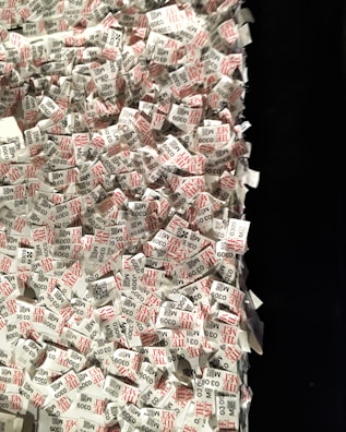 A large pile of printed paper tags, predominantly featuring printed text in black and red. Each tag includes a number and the word 'THE MET', suggesting a connection to a museum or exhibition entry system. The tags are scattered and overlapping each other, creating a sense of chaos and disorganization.