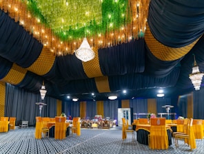 A luxurious indoor banquet hall features a stunning ceiling decorated with lush greenery and sparkling lights. Elegant chandeliers hang from the ceiling, providing a warm glow. The seating area is arranged with vibrant orange chairs around circular tables, all set on an intricately patterned carpet. The walls are adorned with a combination of dark drapery and golden geometric patterns.