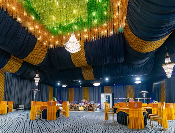 A luxurious indoor banquet hall features a stunning ceiling decorated with lush greenery and sparkling lights. Elegant chandeliers hang from the ceiling, providing a warm glow. The seating area is arranged with vibrant orange chairs around circular tables, all set on an intricately patterned carpet. The walls are adorned with a combination of dark drapery and golden geometric patterns.