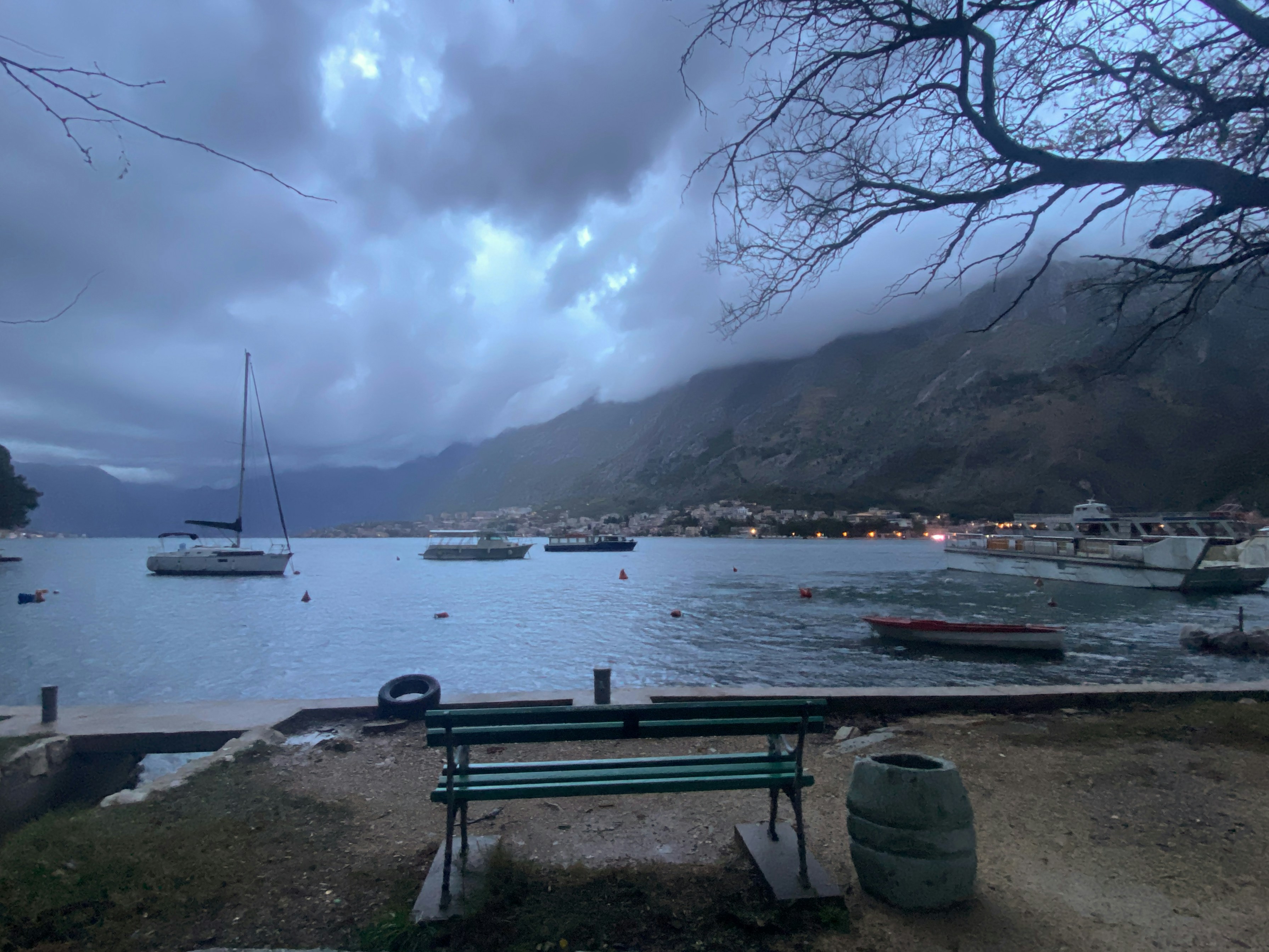 a bench sitting next to a body of water under a cloudy sky, The Bay of Kotor on a cloudy day