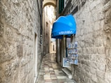 A narrow, cobblestone alleyway lined with textured stone walls. On the right, a small shop with a bright blue awning labeled 'souvenir' displays various postcards and souvenirs. The alleyway is deserted and leads towards an archway in the distance, with more stone buildings visible beyond.