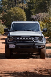 A rugged off-road vehicle parked on a dirt path surrounded by trees, casting a shadow underneath. The front grille displays bold lettering, and the tires have a deep tread pattern suitable for off-road conditions. Sunlight filters through the leaves, illuminating parts of the vehicle.