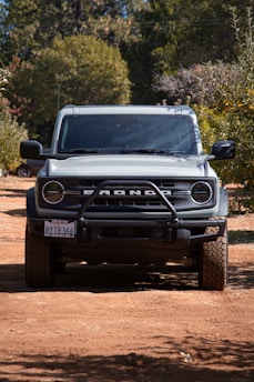 A rugged off-road vehicle parked on a dirt path surrounded by trees, casting a shadow underneath. The front grille displays bold lettering, and the tires have a deep tread pattern suitable for off-road conditions. Sunlight filters through the leaves, illuminating parts of the vehicle.