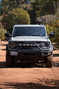 A rugged off-road vehicle parked on a dirt path surrounded by trees, casting a shadow underneath. The front grille displays bold lettering, and the tires have a deep tread pattern suitable for off-road conditions. Sunlight filters through the leaves, illuminating parts of the vehicle.