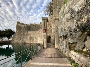 A historical stone fortress stands with tall walls and a walkway leading to an arched entrance. The structure is surrounded by a moat with still blue-green water reflecting part of the fortress. The sky is partly cloudy, and a tree is visible beyond the moat. Metal railings border the walkway for safety. The walls of the fortress display signs of aging and weathering.