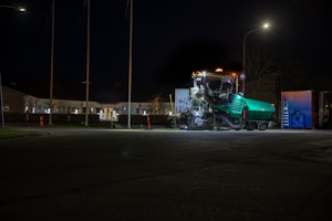 A large industrial machine is illuminated at night, parked in an open area near a building. The machine has multiple components and lights, with a green body and some metallic parts reflecting the ambient light. Nearby is a blue metal structure with a ladder. The setting is dimly lit, with surrounding buildings under a clear, dark sky.