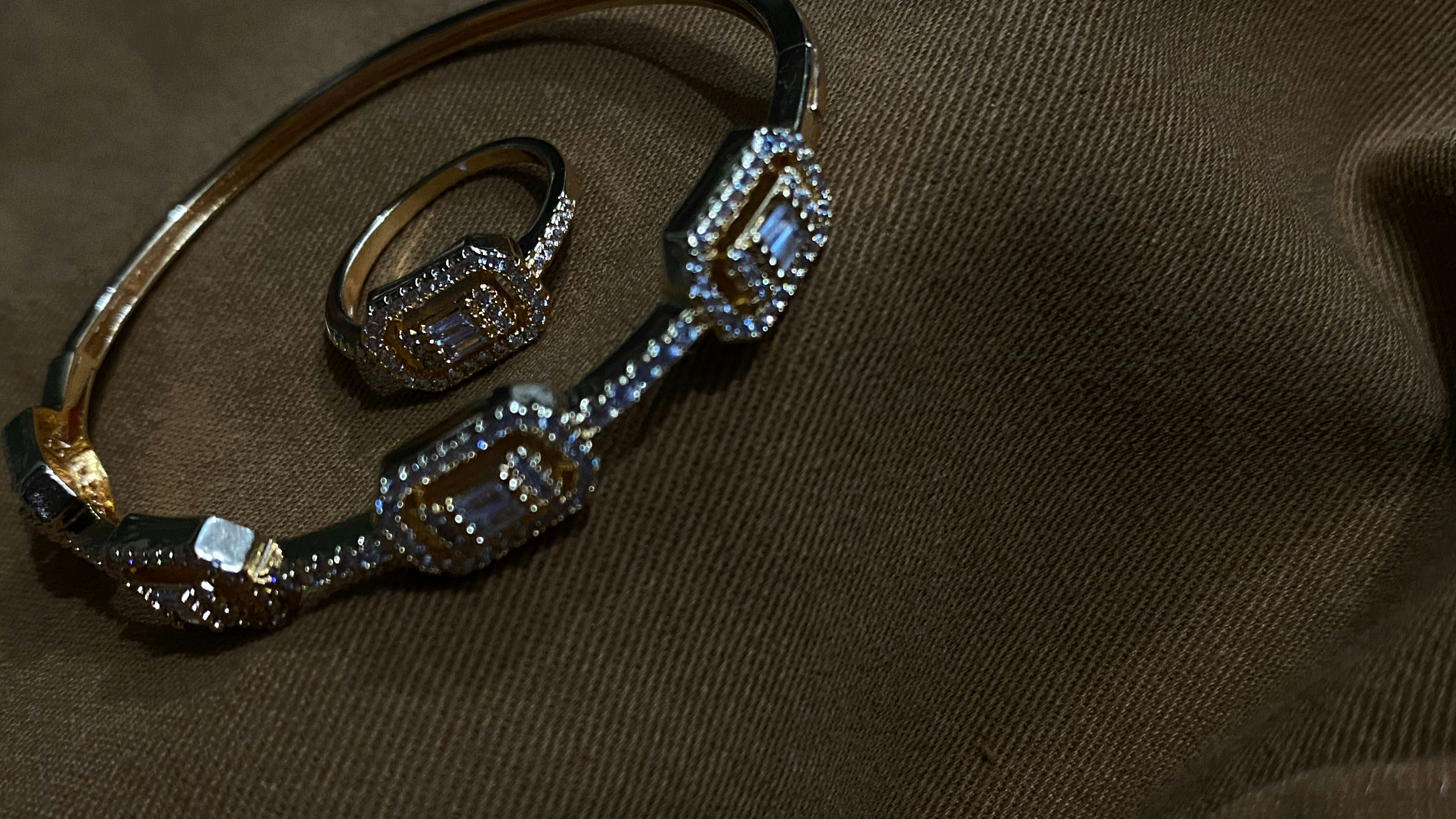 A gold bracelet and ring adorned with intricate geometric patterns and small gemstones are placed on a dark brown fabric with subtle texture.