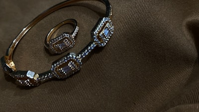 A gold bracelet and ring adorned with intricate geometric patterns and small gemstones are placed on a dark brown fabric with subtle texture.