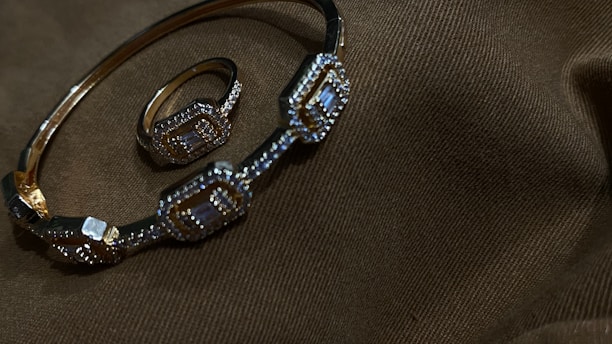 A gold bracelet and ring adorned with intricate geometric patterns and small gemstones are placed on a dark brown fabric with subtle texture.
