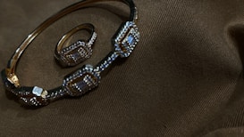 A gold bracelet and ring adorned with intricate geometric patterns and small gemstones are placed on a dark brown fabric with subtle texture.