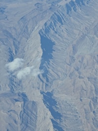 An aerial view of a mountainous landscape with steep ridges and shadowed valleys. The terrain features intricate textures and patterns formed by the geological structure. A few small clouds hover above the mountains, casting soft shadows on the surface.