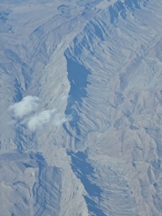 An aerial view of a mountainous landscape with steep ridges and shadowed valleys. The terrain features intricate textures and patterns formed by the geological structure. A few small clouds hover above the mountains, casting soft shadows on the surface.