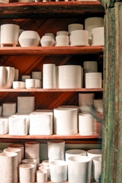 Shelves lined with various ceramic containers, including cups, bowls, and cylindrical vessels in a range of sizes. The pottery is mostly white with some speckled designs, and the wooden shelves provide a warm contrast.