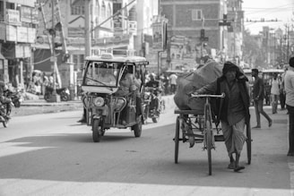 A bustling street scene with a man pushing a loaded cycle rickshaw on a city road. An auto-rickshaw carries passengers in the background, while people walk along the sidewalk. The environment is urban, with buildings, street signs, and a variety of vehicles visible.