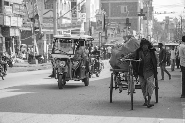 A bustling street scene with a man pushing a loaded cycle rickshaw on a city road. An auto-rickshaw carries passengers in the background, while people walk along the sidewalk. The environment is urban, with buildings, street signs, and a variety of vehicles visible.