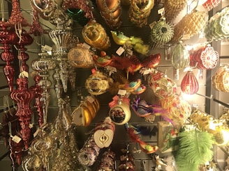 An assortment of colorful and ornate Christmas ornaments hanging on display. The collection features a variety of shapes and designs, including traditional baubles, birds with feathers, and decorative finials. The ornaments are made from materials like glass and metal, adorned with glitter, beads, and intricate patterns.