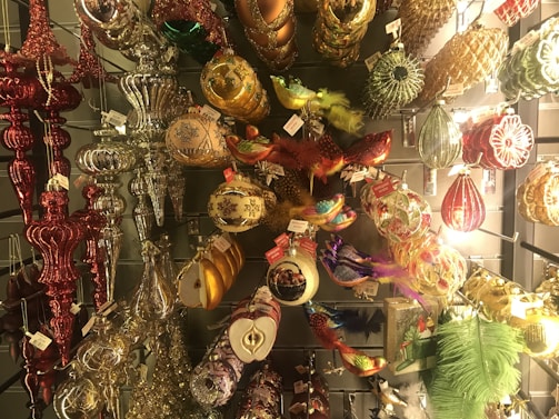 An assortment of colorful and ornate Christmas ornaments hanging on display. The collection features a variety of shapes and designs, including traditional baubles, birds with feathers, and decorative finials. The ornaments are made from materials like glass and metal, adorned with glitter, beads, and intricate patterns.