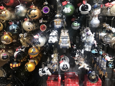 A collection of intricately designed Christmas ornaments featuring popular characters and themes. The ornaments include representations of wizards, stormtroopers, robots, astronauts, and mythical creatures. They are primarily spherical with various decorations and come in a range of colors including gold, black, white, and silver. The display suggests a blend of fantasy and science fiction elements.