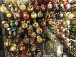 An array of colorful Christmas ornaments displayed on a rack, each featuring intricate designs. The ornaments come in various shapes such as balls, stars, and teardrops, and are decorated with golden, red, and green motifs. Some ornaments are embellished with painted scenes and figures, while others have glittery decorations. Tags are attached to many of the ornaments.