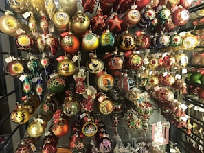 An array of colorful Christmas ornaments displayed on a rack, each featuring intricate designs. The ornaments come in various shapes such as balls, stars, and teardrops, and are decorated with golden, red, and green motifs. Some ornaments are embellished with painted scenes and figures, while others have glittery decorations. Tags are attached to many of the ornaments.