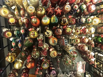 An array of colorful Christmas ornaments displayed on a rack, each featuring intricate designs. The ornaments come in various shapes such as balls, stars, and teardrops, and are decorated with golden, red, and green motifs. Some ornaments are embellished with painted scenes and figures, while others have glittery decorations. Tags are attached to many of the ornaments.