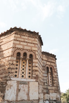 A historic building features intricate brickwork and arched windows, with ornate stone detailing. The structure has a weathered appearance, suggesting age and historical significance. The architecture reflects traditional design elements, including patterned bricks and detailed window casings.