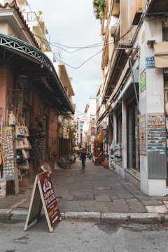 A narrow street flanked by old buildings with signs, shops, and decorations. The path is paved with stone, and a person walks in the distance. There's a sign for 'Jaipur Palace' indicating Halal food, along with various other items displayed outside the shops.