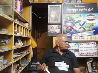 An older man with short, gray hair sits in a small, cluttered shop. He wears a black shirt with playing card designs and holds a cigarette. Behind him, the shop walls are covered with various advertisements, including one for cigarettes and another featuring bold graphics. Shelves to the left are stacked with various goods, including snacks and a QR code display for digital payments.