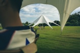 A large, triangular white tent set up on a sprawling green lawn under a partly cloudy sky. The scene includes outdoor furniture like chairs and tables, with some string lights hanging along the edges of the tent. Trees and bushes can be seen in the background.