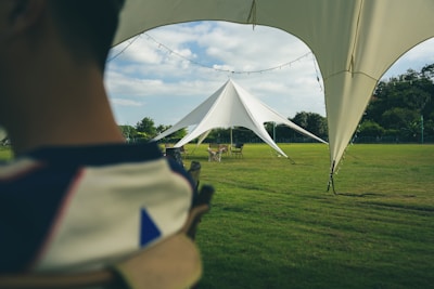 A large, triangular white tent set up on a sprawling green lawn under a partly cloudy sky. The scene includes outdoor furniture like chairs and tables, with some string lights hanging along the edges of the tent. Trees and bushes can be seen in the background.