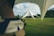 A large, triangular white tent set up on a sprawling green lawn under a partly cloudy sky. The scene includes outdoor furniture like chairs and tables, with some string lights hanging along the edges of the tent. Trees and bushes can be seen in the background.