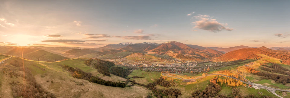 The image presents a panoramic view of a mountainous landscape, captured from an aerial perspective. The mountains, bathed in warm hues of orange and yellow, rise majestically against the backdrop of a clear blue sky dotted with fluffy white clouds. The sun is setting behind these towering peaks, casting long shadows that stretch across the scene. In the foreground, a lush green valley unfolds, teeming with trees and vegetation. The image is devoid of any human presence or text, allowing for a sense of tranquility and solitude to envelop the viewer.