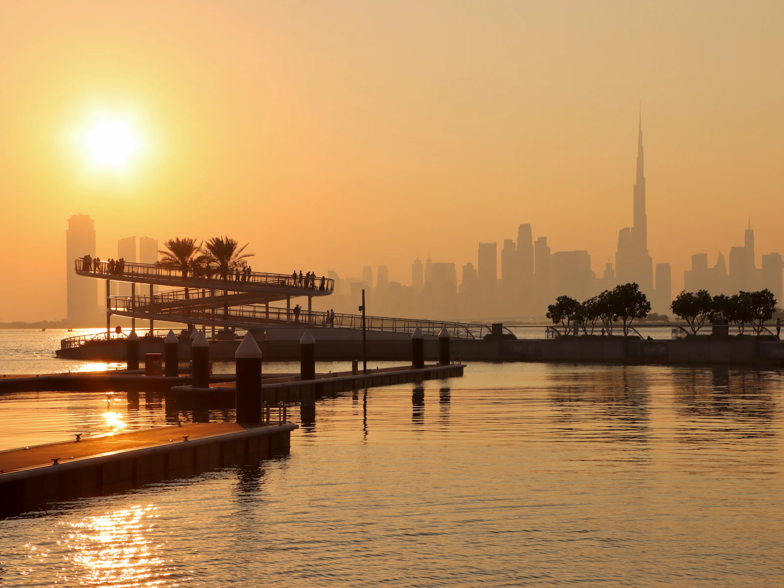 The curving elevated viewing boardwalk and floating marina pontoons at Dubai Creek Harbour at sunset, Downtown Dubai and Burj Khalifa in the distance